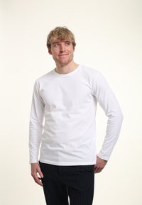 Bílé tričko s dlouhým rukávem a kulatým výstřihem, vyrobené z měkké tkaniny. Nosené s černými pruhovanými kalhotami, tričko má hladkou texturu.