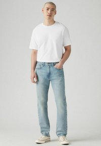 Weißes T-Shirt mit kurzen Ärmeln, hellblaue gerade Jeans, trägt helle Sneakers, steht vor einem neutralen Hintergrund.