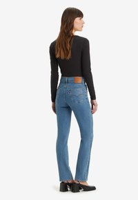 Jeans azuis de cintura alta com bolsos traseiros com detalhes de tachas, combinados com uma blusa preta de manga longa e sapatos de salto pretos.