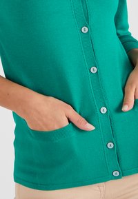 Cardigan verde con chiusura a bottoni, polsini a costine e tasca sul lato sinistro. Realizzato in un tessuto a maglia morbido.