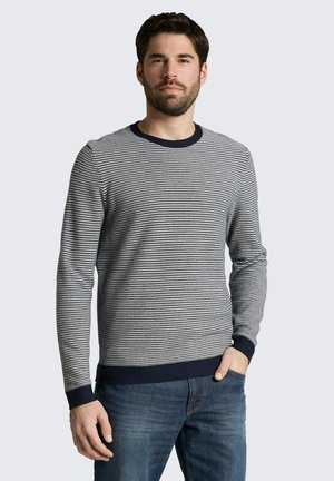 Homme aux cheveux foncés et à la barbe, portant un pull ajusté à manches longues à rayures horizontales et un jean bleu, debout avec une main dans la poche.