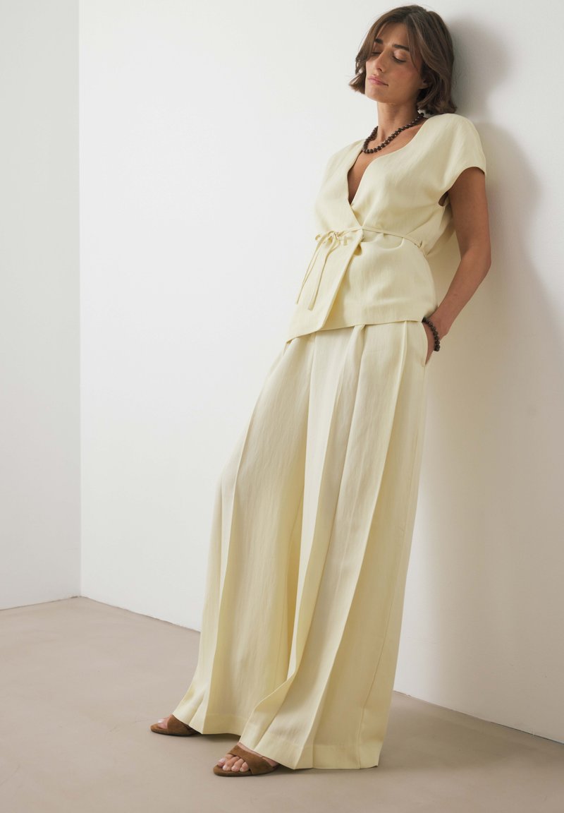 Sieviete balstās pret sienu, uzvilkusi krēmkrāsas platās bikses, saskaņotu īso piedurkņu krekliņu, brūnu kaklarotu, aproci un sandales.