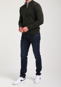 Pull-over vert foncé avec un motif en losange texturé, col avec demi-fermeture éclair, porté sur une chemise blanche à motifs, associé à un jean denim foncé et des baskets blanches.
