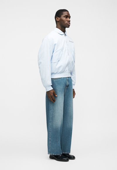 Homme debout portant une veste bomber bleu clair, un jean bleu délavé à jambes larges et des chaussures en cuir noir, sur un fond blanc uni.