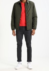 Man i olivgrön knäppjacka över en röd tröja, svarta byxor, vita strumpor och vita sneakers, som står mot en vit bakgrund.