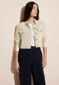 Krämfärgad denimjacka med silverfärgade knappar, två bröstfickor, kort design och åtsittande ärmar, kombinerad med en vit topp och marinblå byxor.