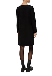 Czarna sukienka z długim rękawem o teksturowanym wzorze i prostym kroju, sięgająca do połowy uda, w połączeniu z czarnymi botkami sięgającymi do kolan.