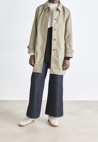 Trench-coat beige clair à boutons, associé à un pull bicolore noir et crème et à un pantalon large en denim foncé. Des baskets blanches complètent le look.