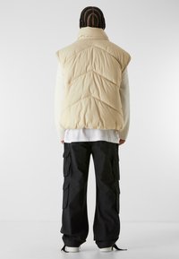 Beige padded vest with a high collar and chevron stitching, worn over a white long-sleeve shirt and paired with black cargo pants.