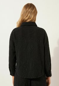 Giacca nera con motivo floreale testurizzato, maniche lunghe e colletto. Presenta polsini con bottoni e un orlo dritto sul retro.