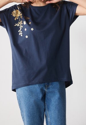 Persona que lleva una camisa azul marino con diseño floral dorado en el hombro y vaqueros azules, manos cerca de la cara, recortado a la altura de la barbilla y las caderas.