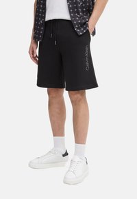 Pantalones cortos de algodón negros con cintura elástica y cordón ajustable, con un estampado de logotipo en el lateral. Combinados con zapatillas blancas y calcetines.
