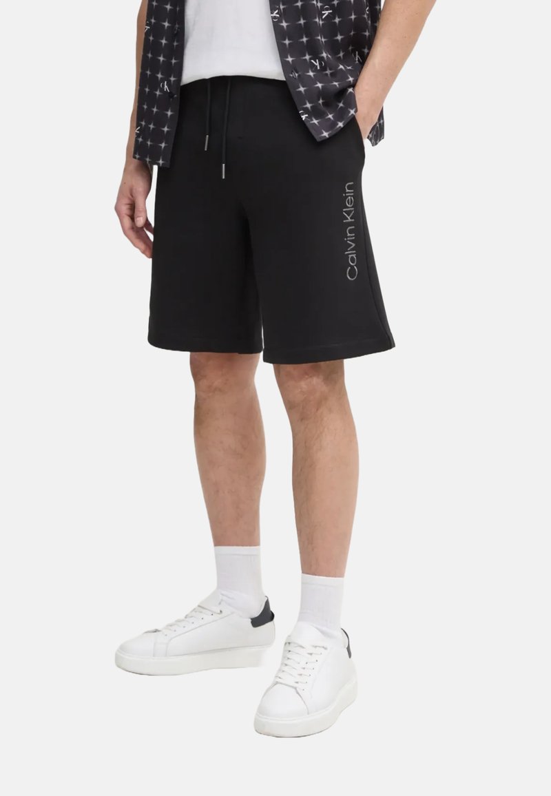 Pantalones cortos de algodón negros con cintura elástica y cordón ajustable, con un estampado de logotipo en el lateral. Combinados con zapatillas blancas y calcetines.