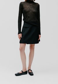 Schwarzes, langärmliges, durchsichtiges Oberteil, tailliert geschnitten, kombiniert mit einem schwarzen A-Linien-Rock mit einer flachen Tasche. Schwarze flache Schuhe mit einer Schleifendetail.