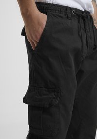 Person wearing black cargo shorts with elastic waistband and drawstring, hand resting inside front pocket, visible tattoo on arm.
