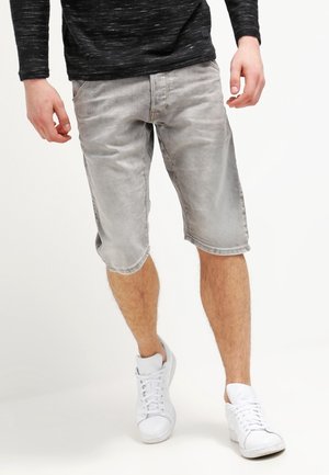 Homme portant un short gris délavé jusqu'aux genoux, un haut à manches longues noir et des baskets blanches, marchant devant un fond uni.