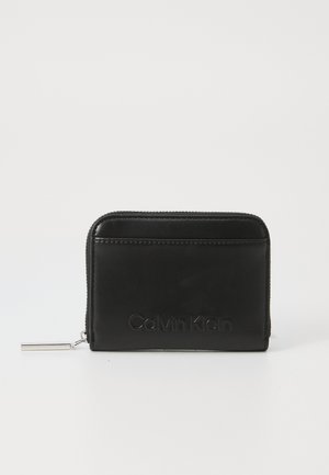 Portofel din piele neagră cu închidere prin fermoar. Prezintă logo-ul embosat "Calvin Klein" pe partea din față. Textură netedă și formă rectangulară.