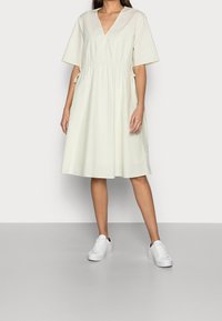 Vaaleankeltainen puuvillamekko, jossa on V-mallinen kaula-aukko, lyhyet hihat ja elastinen vyötärö, yhdistettynä valkoisiin lenkkareihin harmaalla pinnalla.