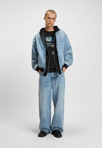 Chaqueta de denim con un lavado azul claro, sudadera negra debajo, camiseta gráfica, vaqueros de pierna ancha y zapatos negros.