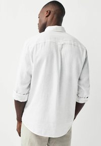 Uomo di spalle che indossa una camicia bianca a maniche lunghe con le maniche arrotolate e pantaloni chiari, di fronte a uno sfondo semplice e chiaro.