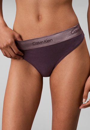 Kvinde iført lilla Calvin Klein undertøj, der holder i linningen på den ene side og viser maven og den øverste del af lårene.