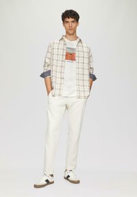 Geruite langarmsgem shirt in beige en bruin over een wit graphic t-shirt; gecombineerd met witte broek en witte sneakers met bruine accenten.