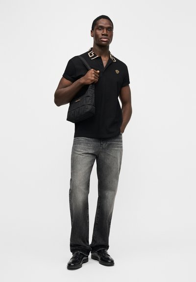 Jaunas vyras, vilkintis juodą polo marškinėlį su raštuota apykakle ir emblemą, blyškiai pilkus džinsus, juodus batus, laikantis juodą siuvinėtą pečių krepšį.