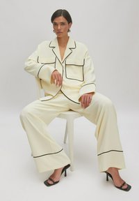 Crèmewit pak met zwarte piping, bestaande uit een losvallende blazer en wijde broekspijpen. Model in strappy zwarte hakken, zittend op een stoel.