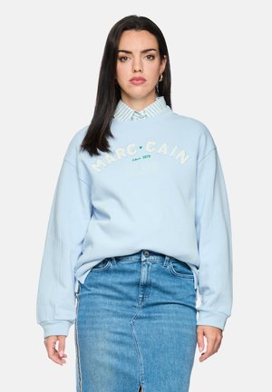 Frau mit langem dunklem Haar, die einen hellblauen Marc Cain Sweatshirt über einem gestreiften Hemdkragen und einem blauen Jeansrock trägt, stehend vor einem schlichten Hintergrund.