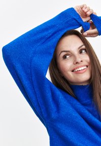 Niebieski sweter dzianinowy z wysokim kołnierzem i długimi, luźnymi rękawami. Posiada teksturowane wykończenie i ściągacze. 