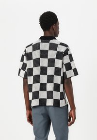 Chemise à manches courtes avec un motif à carreaux noir et blanc, fabriquée en tissu texturé, avec un col noir et une coupe décontractée.