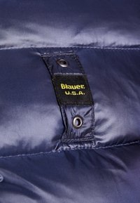 Giacca puffer blu navy in tessuto morbido, con etichetta nera con testo giallo. Include dettagli in metallo e cuciture.