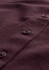 Burgunderfarbener strukturierter Stoff mit sichtbarem Knopfdetail, das ein dezentes Rippmuster und glänzende, dunkle Knöpfe entlang der Knopfleiste aufweist.