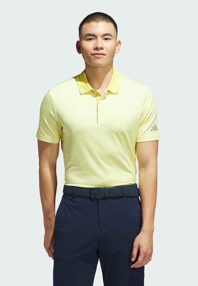 Camiseta polo de color amarillo claro con un patrón texturizado, de manga corta y con cuello. Combinada con pantalones azul marino oscuros y un cinturón texturizado azul marino.