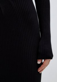 Schwarzes, geripptes Strickkleid mit langen Ärmeln, das eine strukturierte Oberfläche und eine figurbetonte Silhouette aufweist. Es zeigt ein Detail am Handgelenk und eine Hand.