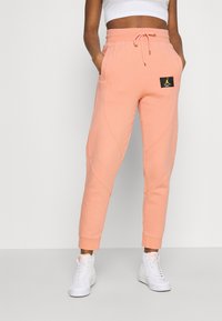 Peach-colored joggers with a drawstring waist, side pockets, tapered leg, and a black logo patch. Made of soft, cozy fabric.