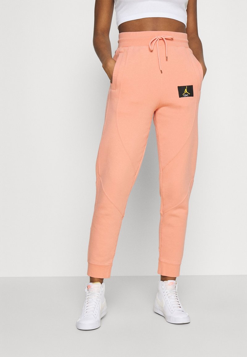 Peach-colored joggers with a drawstring waist, side pockets, tapered leg, and a black logo patch. Made of soft, cozy fabric.