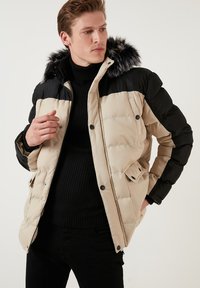 Giacca a vento beige e nera, con cappuccio in pelliccia sintetica, texture trapuntata, chiusura a pressione e tasche laterali. Indossata sopra un maglione nero.