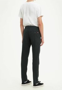 Pantaloni neri in cotone con vestibilità slim, dotati di una tasca posteriore e un logo rosso discreto, abbinati a una maglietta bianca semplice e sneakers nere.