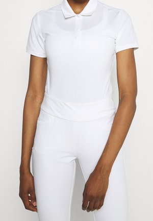 Osoba ubrana w dopasowaną, białą koszulkę polo z krótkim rękawem i białe spodnie z wysokim stanem, stojąca z ramionami swobodnie opuszczonymi wzdłuż ciała.