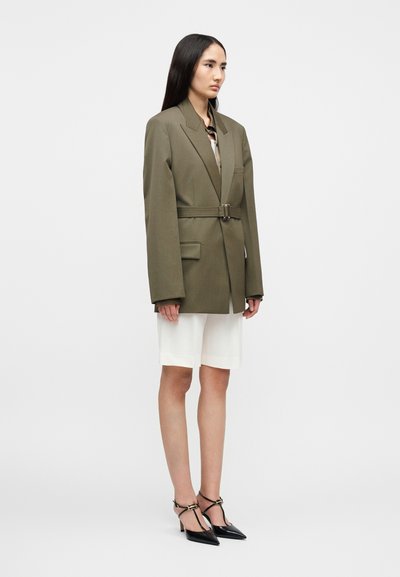 Υπερμεγέθη σακάκι σε ελιάς πράσινο με ζώνη, με δύο μπροστινές τσέπες, φορεμένο πάνω από ανοιχτό κρεμ σορτς, συνδυασμένο με μαύρες μυτερές γόβες.