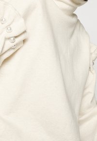 Krémový pulovr s jemnou texturou, který má volány na rukávech zdobené dekorativními perlovými detaily podél ramenního švu.