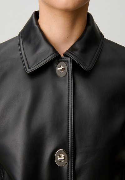 Chaqueta de cuero negra con un cuello ancho y dos grandes botones de palanca plateados abrochados en el pecho.