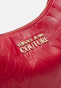 Červená kožená kabelka má embosované vzory, zlatý zip a zlaté logo s nápisem "Versace Jeans Couture" výrazně umístěné.