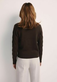 Ciemnobrązowy sweter dzianinowy z detalami splotu w rękawach i ściągaczem na dole, zestawiony z jasnymi spodniami.