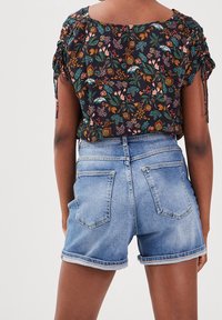 Vista trasera de una persona que lleva una blusa de estampado floral con mangas fruncidas y pantalones cortos de mezclilla azul claro con dobladillo vuelto.