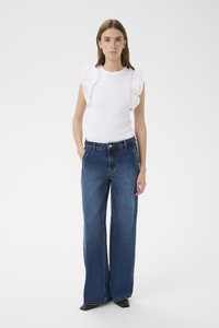 Biely tielkový top s volánikovým detailom na ramenách, v kombinácii so širokými tmavomodrými džínsami, na čistom bielom pozadí.