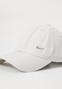 Fehér baseball sapka varrott panelekből és ívelt szélekkel, a bal elülső oldalon egy kis fémes Nike swoosh logóval.