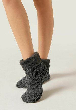 Jambes portant des chaussons en tricot gris foncé avec des bords en fausse fourrure sur un fond clair.
