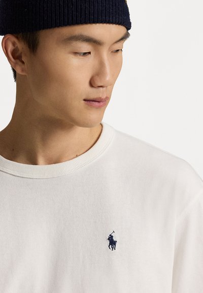 Balta kokvilnas sporta krekls ar ribotātu apkakli un tumši zilu izšūtu polo spēlētāja logo kreisajā krūtī. Redzama mīksta tekstūra.